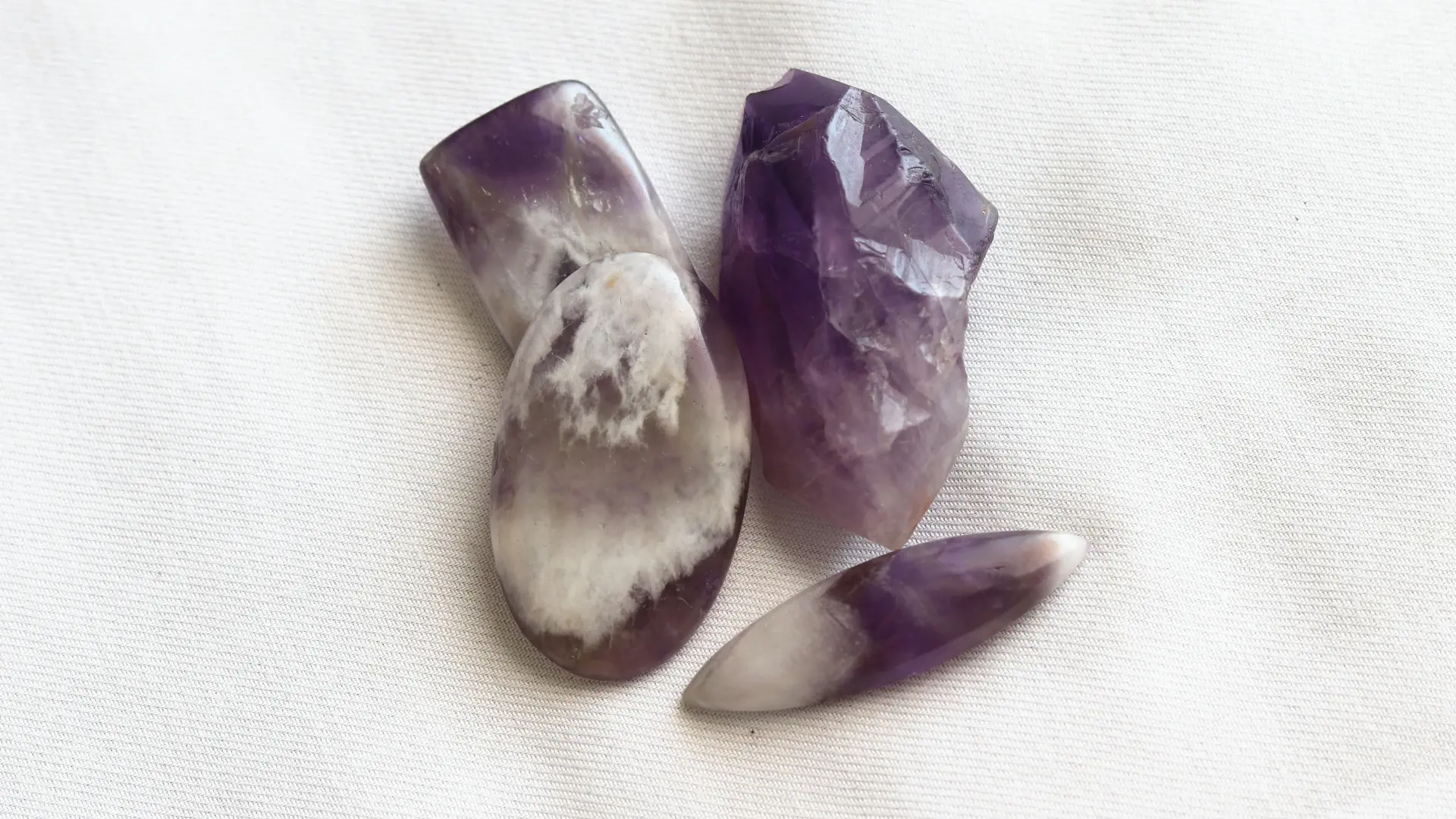 Four amethyst stones on white fabric: a dark purple rough piece and three polished stones banded in purple and white