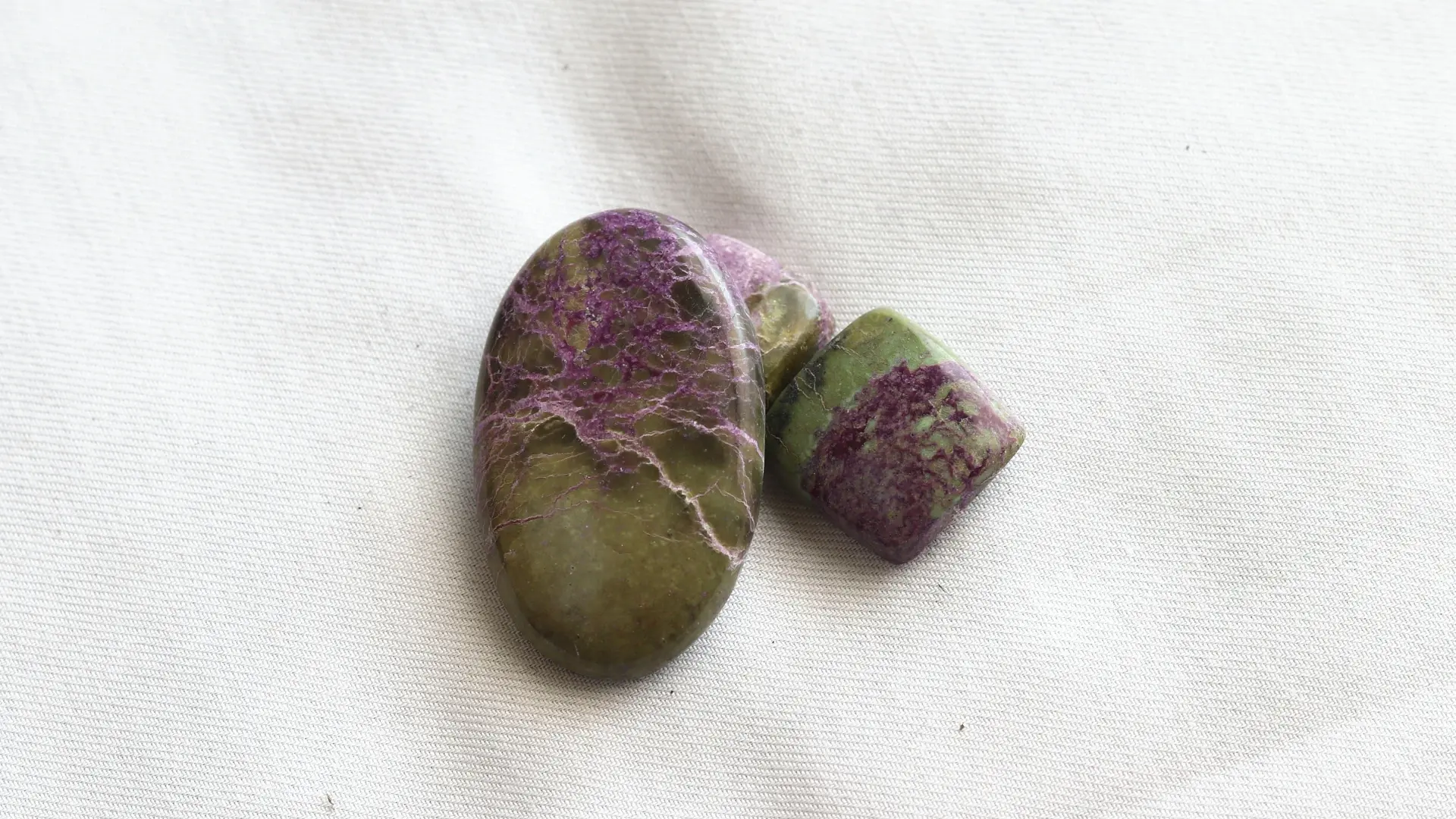 Three polished olive-green atlantisite stones with purple veins and patches, on white fabric