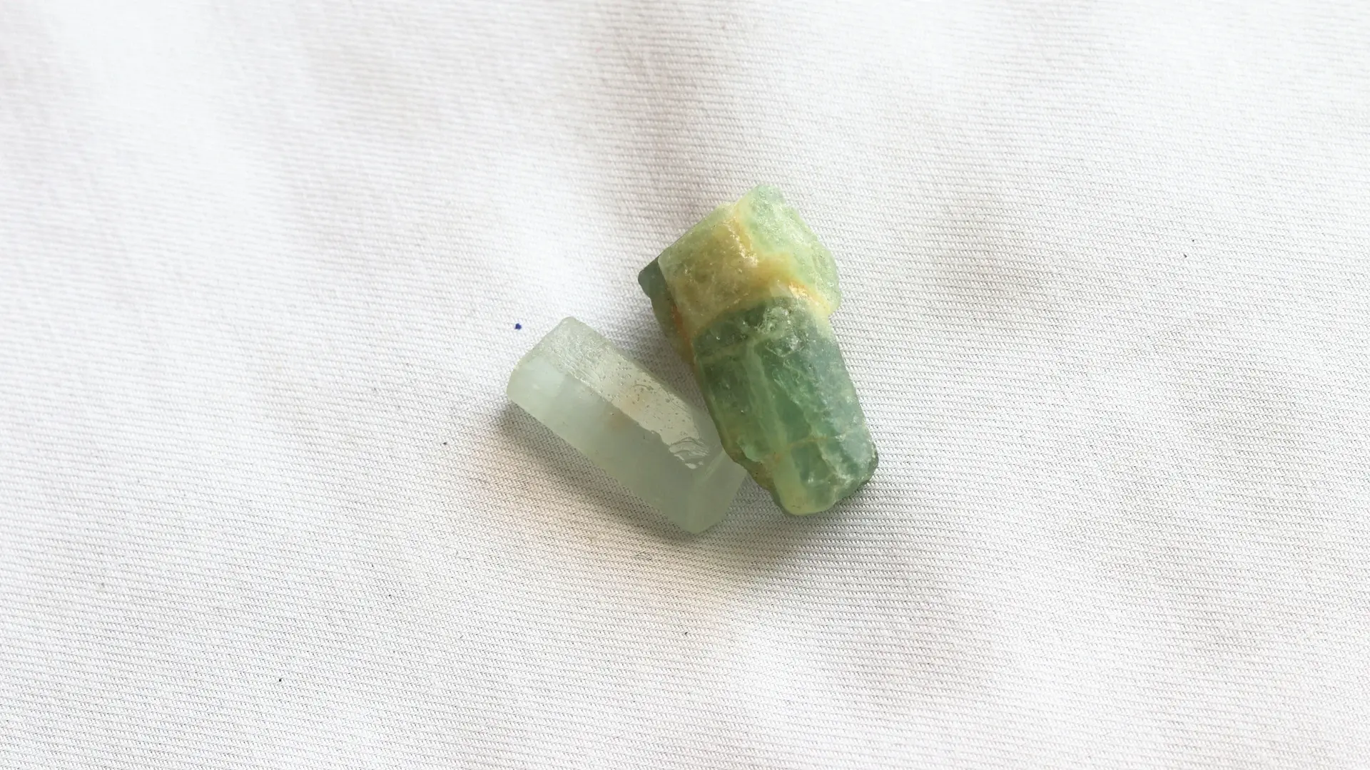 Two raw prismatic aquamarine crystals in pale green-blue, on white fabric