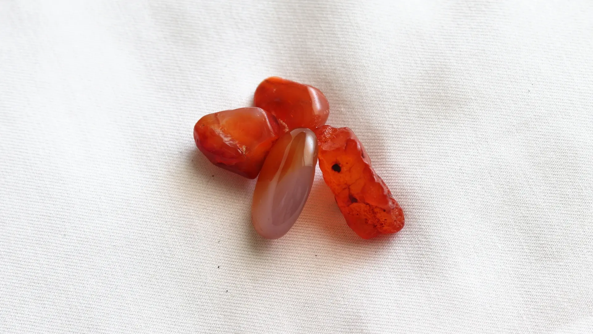 Four orange-red carnelian stones, some polished and some rough, on white fabric