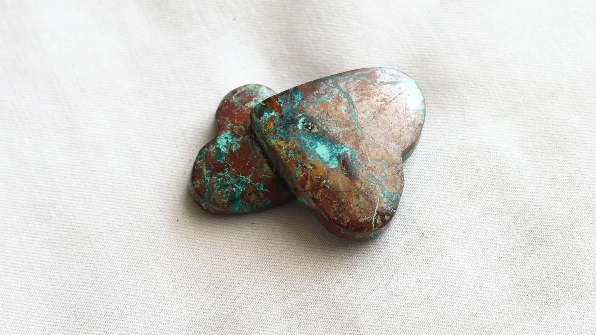 Two polished heart-shaped stones in turquoise, red-brown and gold tones, on white fabric